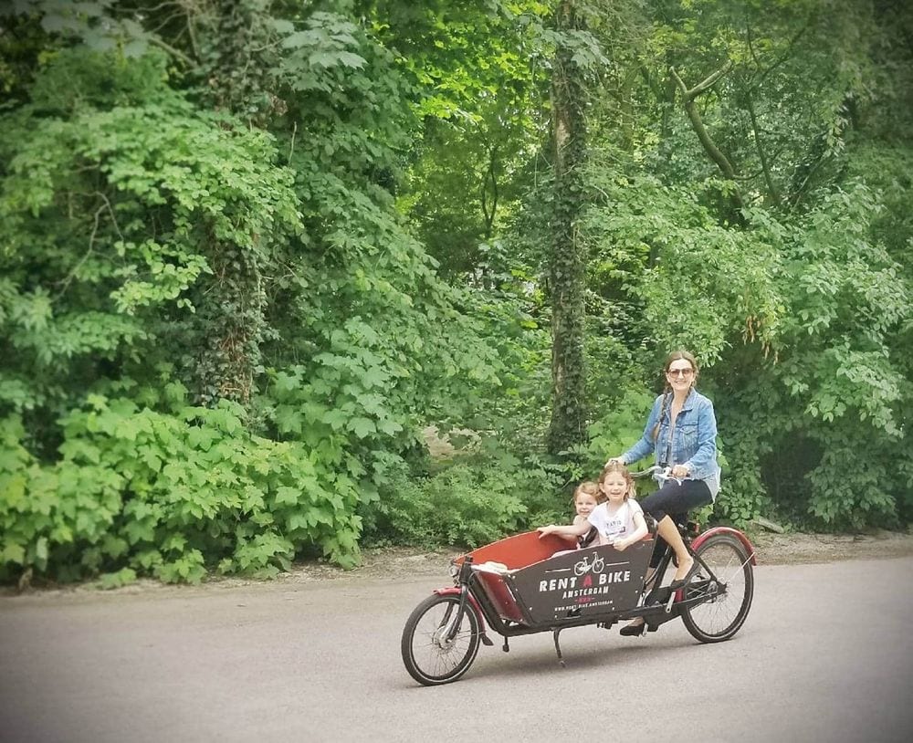 A mother sits atop a bike with a front buggie, which holds her two young girls. All look at the camera with huge smiles. Filling your child's life with experiences is one of the benefits of traveling with kids.