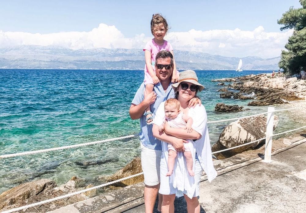 A family of four stands smiling off the coast of a large body of water in Croatia, with mountains on the other shore. The mom holds a baby, while the dad has the older child perched on his shoulders.